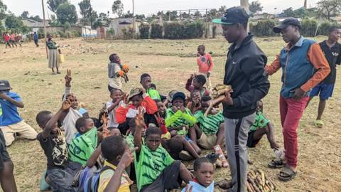 A coach holding sugarcane stands among boys sitting on the pitch, many making peace signs