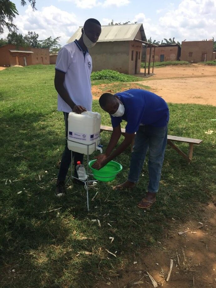Two men demonstrating a jerrycan handwashing station in the settlement