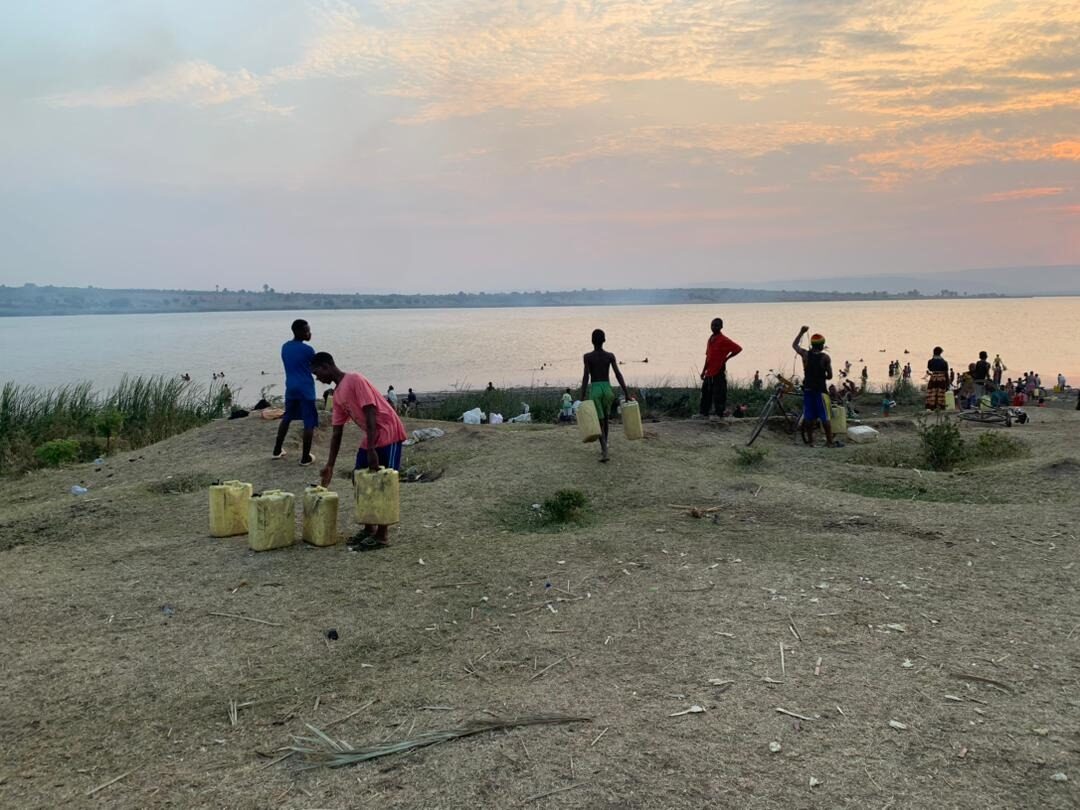 Young people carrying yellow water jerrycans along the lakeshore at sunset near Nakivale