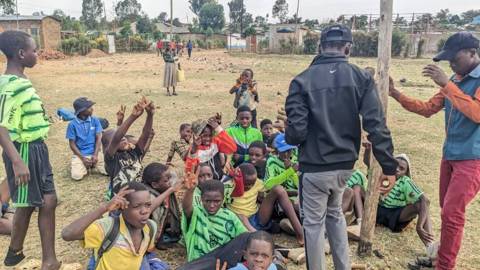 Boys sitting on the pitch raising their hands, seen from behind two coaches