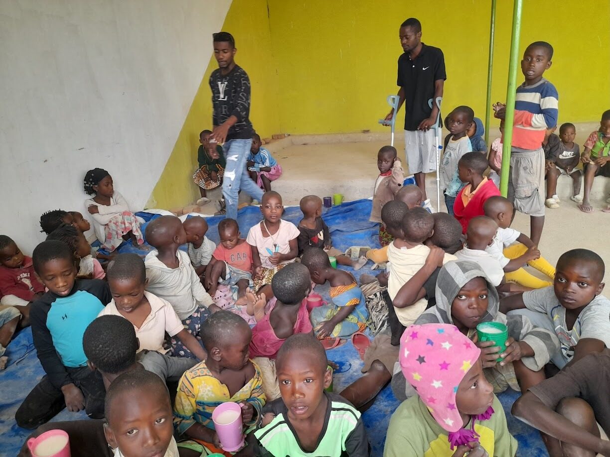 Dozens of children seated on tarpaulins sharing warm porridge at a feeding program