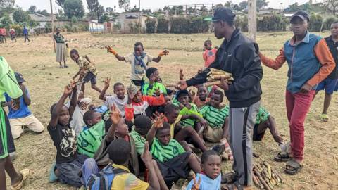 Boys in green shirts sitting on the grass while a coach hands out sugarcane