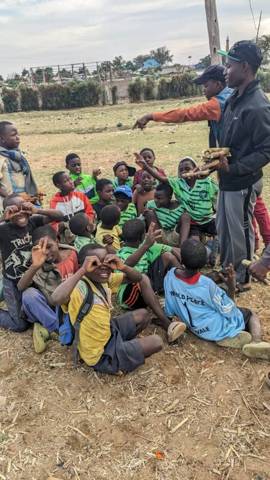 Boys sitting on the ground listening to a coach, some making glasses with their fingers
